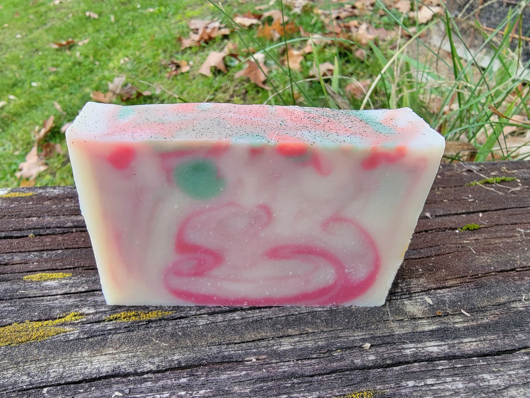White soap with red and green swirls sitting on a ledge with grass and leaves in the background. Glitter is on top of the soap.