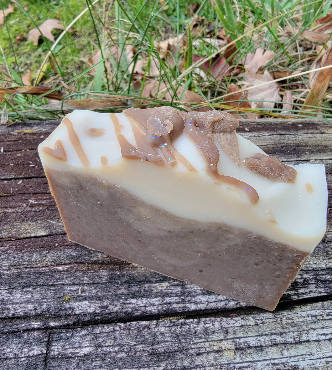 Chocolate colored soap with a white top and chocolate drizzle with glitter sitting on a ledge with grass and leaves in the background.